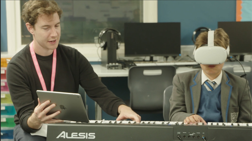 A male teacher, instructing a young boy wearing a VR headset, playing a keyboard, in a classroom setting.